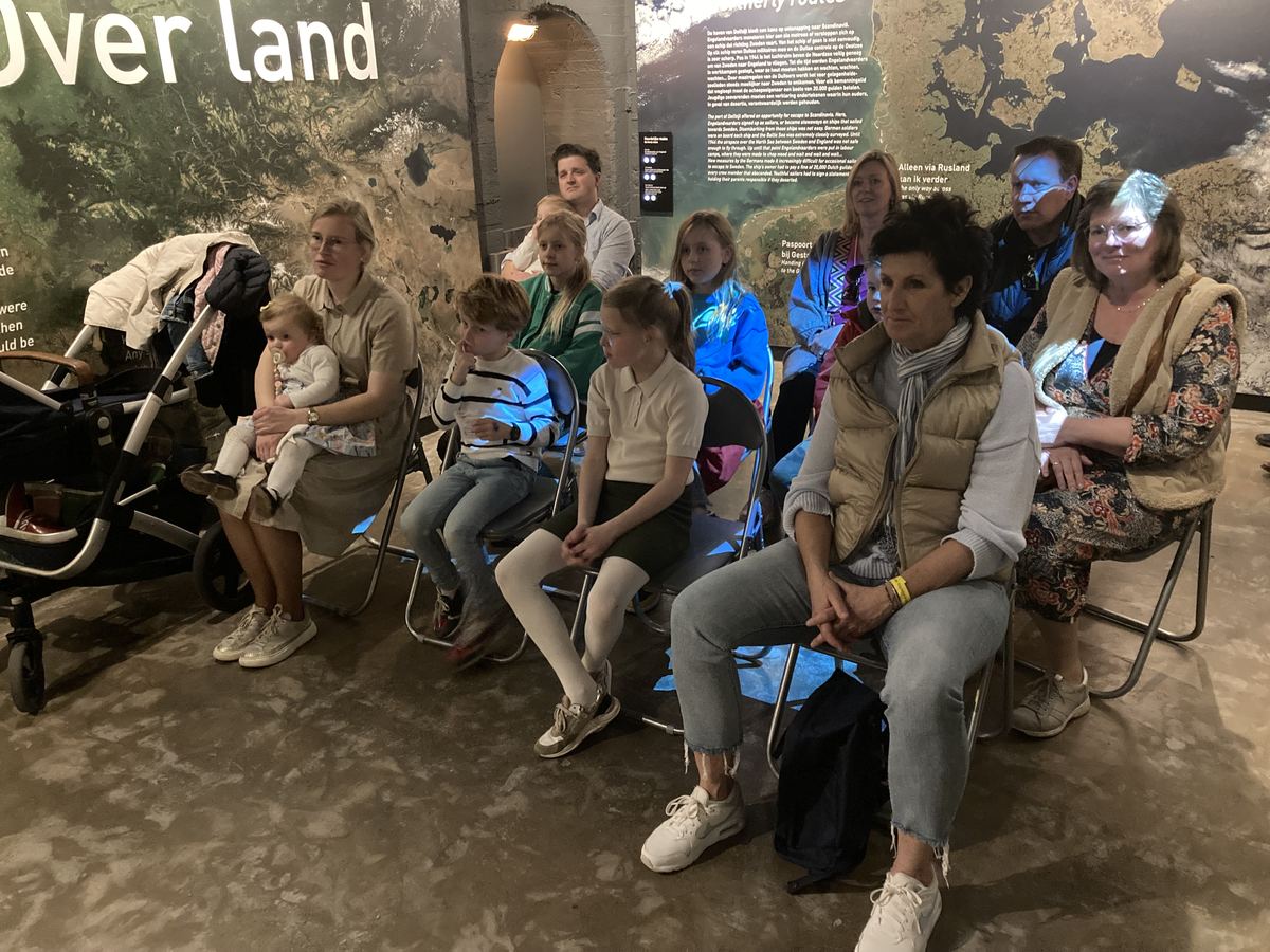 Volle rij bezoekers, van jonge kinderen tot ouderen, op stoelen in de museumzaal