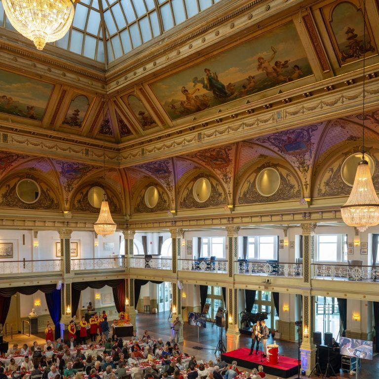De zaal van het Kurhaus in Scheveningen met kroonluchters en beschilderd plafond, vol gasten aan tafel