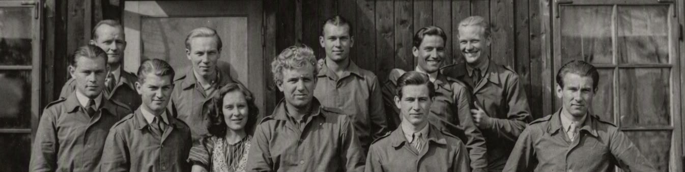 Groepsportret van Engelandvaarders in uniform voor een houten barak, met een vrouw in hun midden