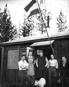 Henk Brandenburg (uiterst links staand) tijdens zijn werkzaamheden als bosbouwer in het gehucht Koppslahyttan, in afwachting van het moment waarop hij vanuit Zweden de oversteek naar Engeland kon maken. Op de foto zijn rechts van hem ook zijn mede-Engelandvaarder Jan (“Puck”) van der Weyden, Frans Pauli en Memco Mulder te zien (november 1943). Vaderlandse driekleur in top boven de blokhut van de naar Zweden uitgeweken Engelandvaarders (Koppslahyttan)